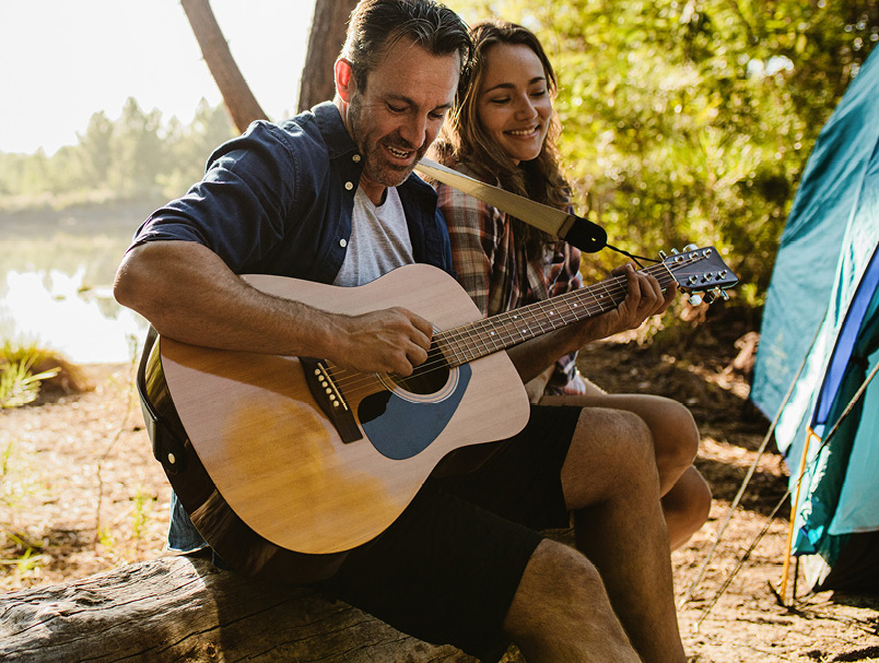 A man and woman in their late 30s/early 40s are hanging out at their campsite located next to a lake. The man plays the guitar as the woman listens, smiling.